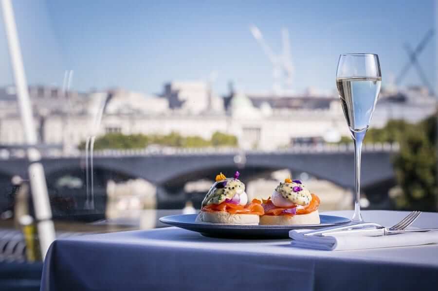A glass of champagne and a plate of sandwiches on a table overlooking the river thames.