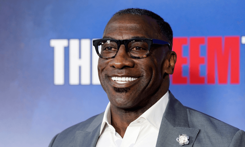 A black man wearing glasses and a suit smiles at the red carpet.