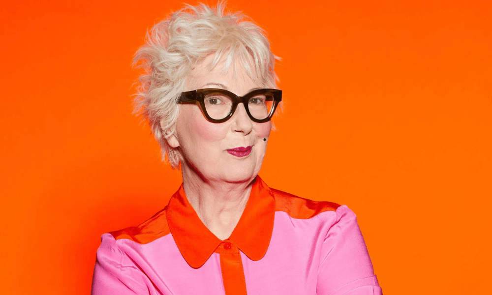 A woman with glasses and a pink shirt is posing in front of an orange background.