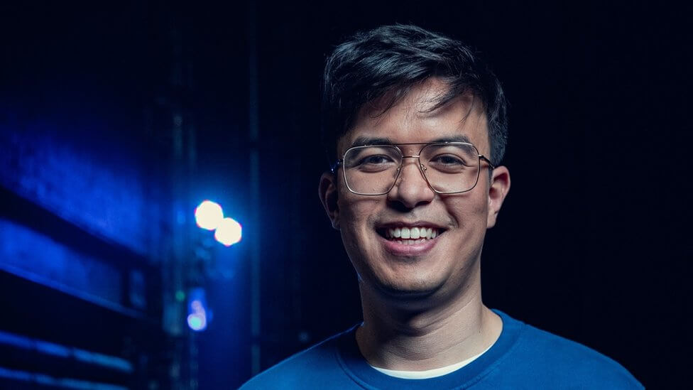 A smiling young man with glasses, wearing a blue shirt, in front of a dark, blurred background with a blue stage light.