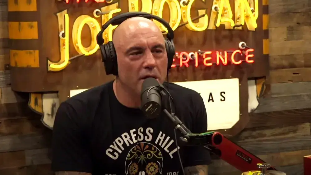Man speaking into a microphone on a podcast set with a sign that reads "joe rogan experience" in the background.