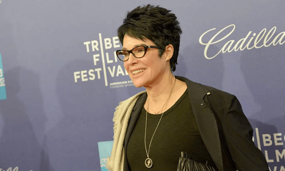A woman with short black hair, wearing glasses, a black top, and a necklace, smiles as she poses on the red carpet at the tribeca film festival.