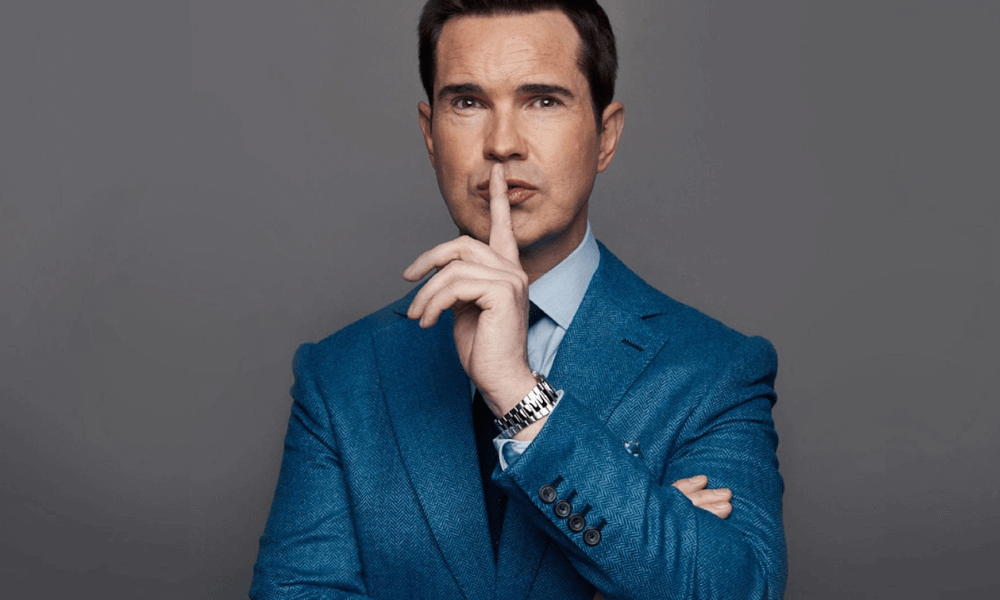 A man in a blue suit making a silence gesture with his finger to his lips.