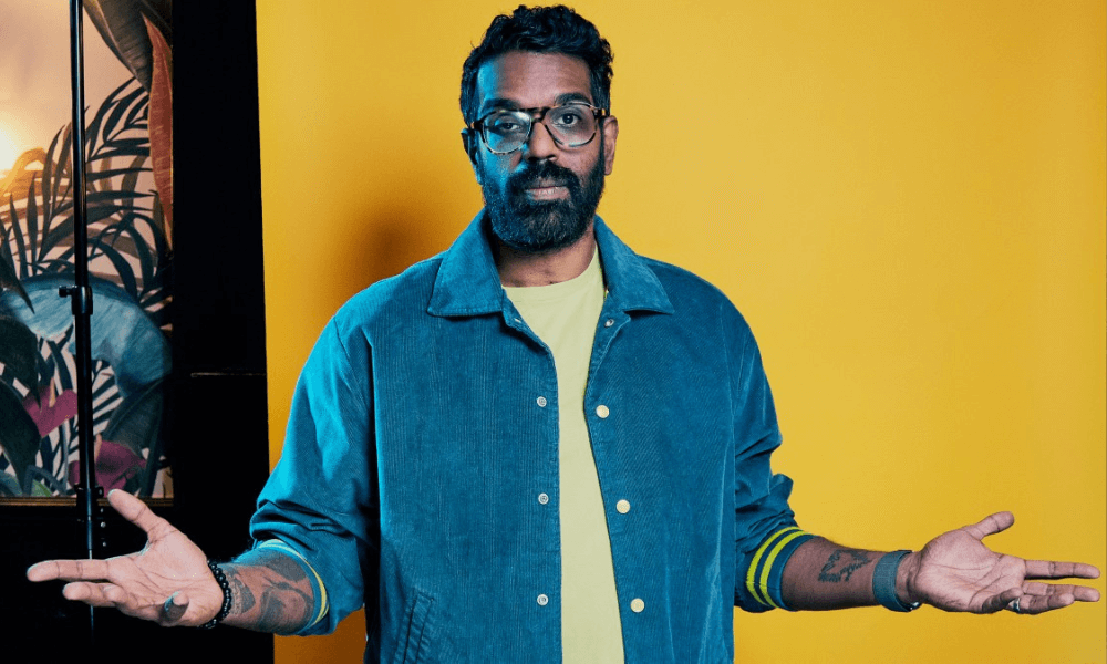 A man with glasses and a beard standing in front of a yellow wall, with an open denim shirt over a teal t-shirt, looking slightly perplexed with his palms facing up.