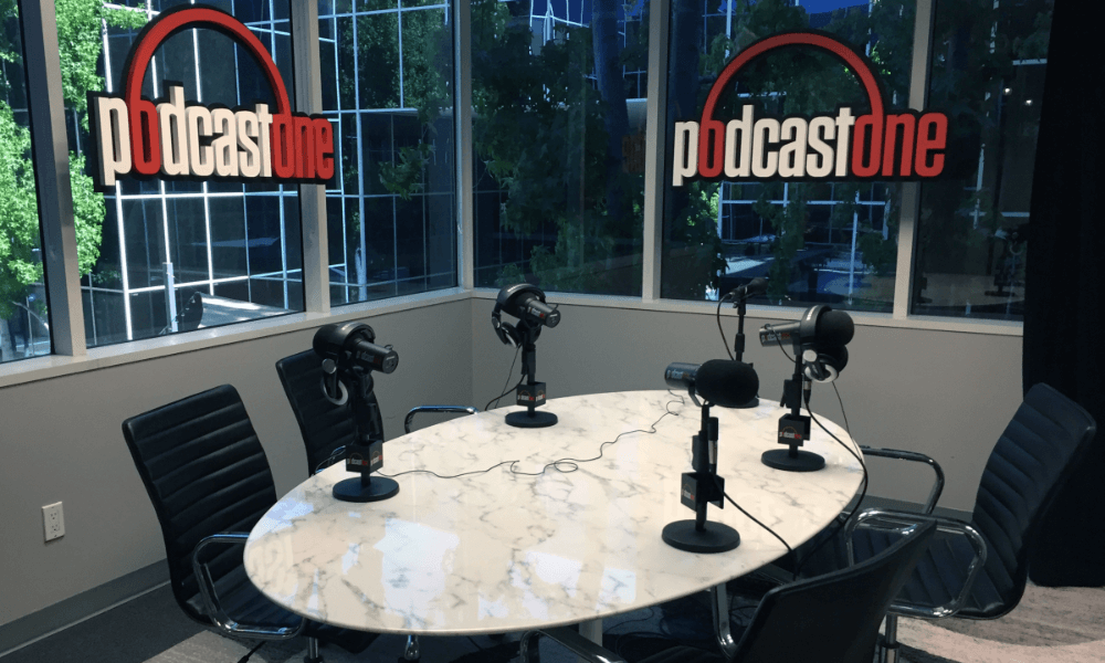 A podcast recording studio with microphones and chairs, featuring the logo "podcastone" on the window.