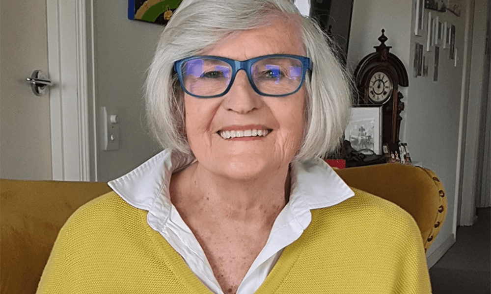 A smiling elderly woman with gray hair wearing blue-framed glasses, a yellow sweater, and a white collared shirt, sitting indoors.