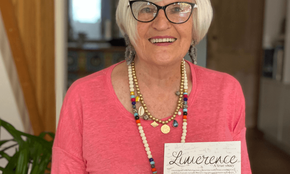 An elderly woman with glasses smiling while holding a book titled "limerence.