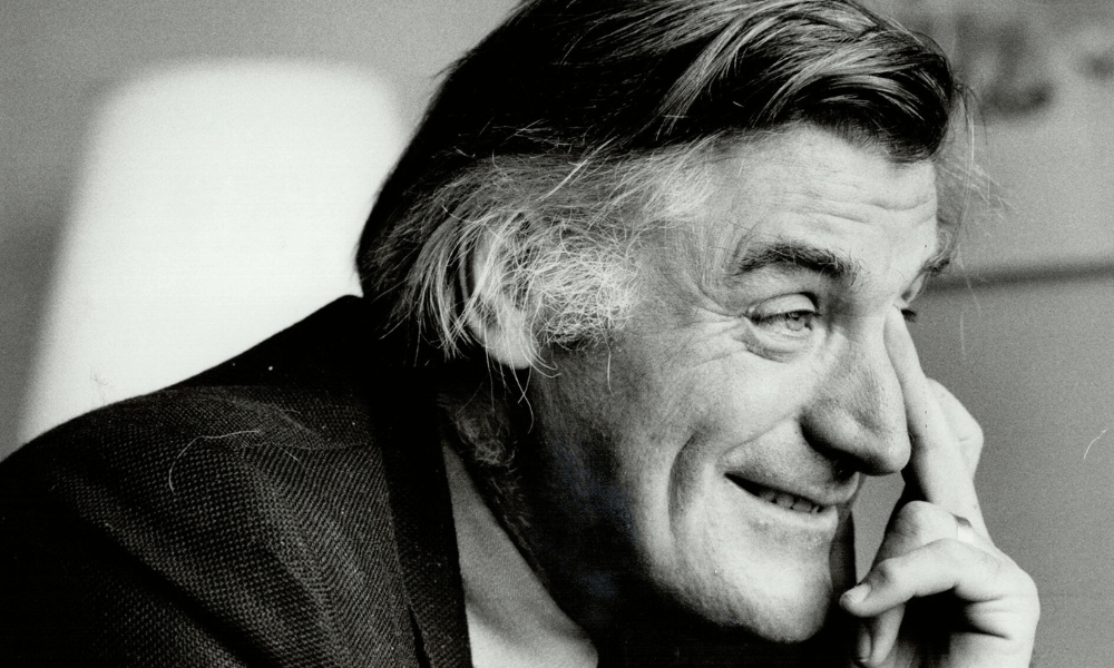 Black and white photo of a smiling man with combed-back hair resting his chin on his hand.