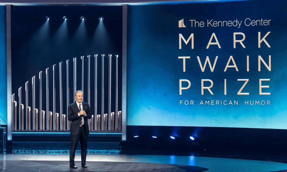 A man in a suit stands on a stage beside a screen displaying "the kennedy center mark twain prize for american humor.