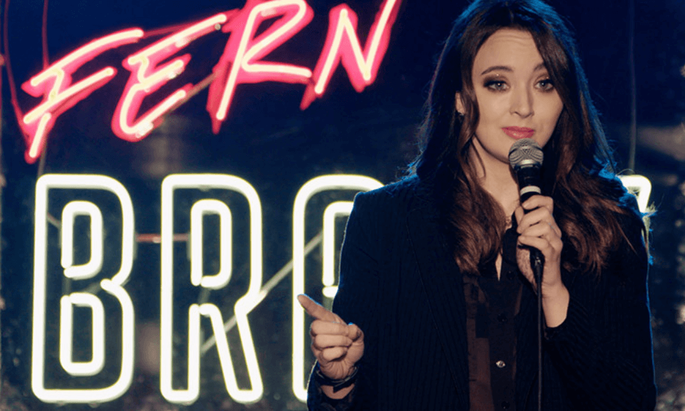 A woman holding a microphone stands in front of a neon sign while addressing an audience.