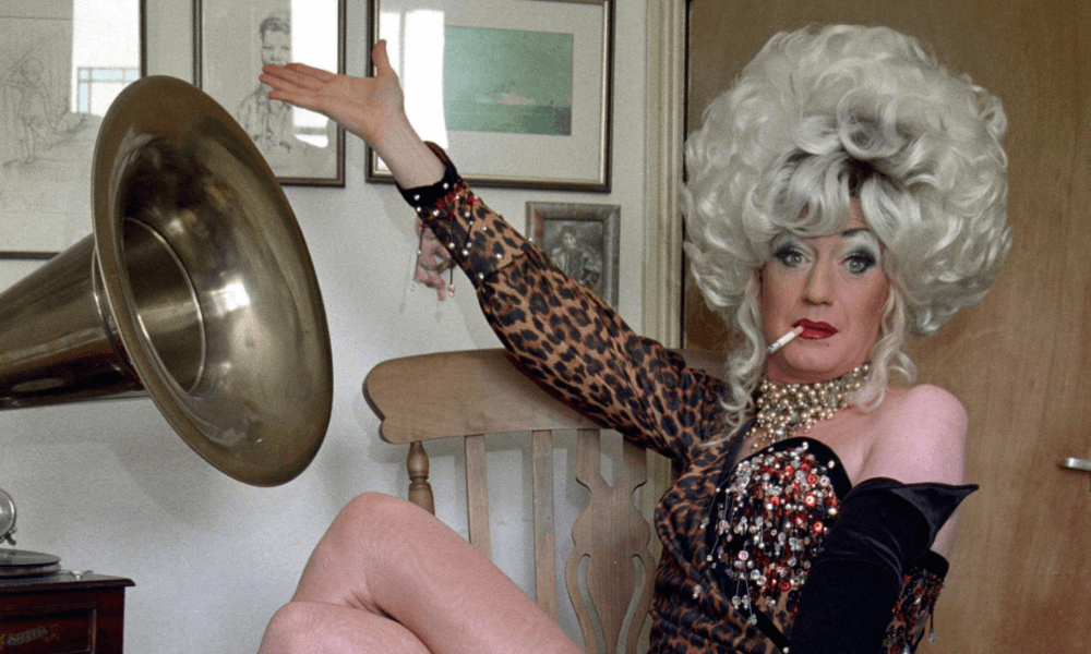 Person in flamboyant drag costume with a cigarette posing by a gramophone.
