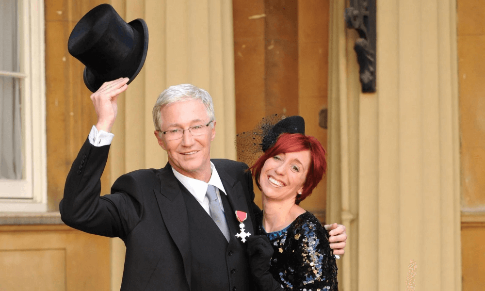 A man in a suit holding a top hat aloft and a smiling woman in a black dress with a fascinator standing together outside a formal building.