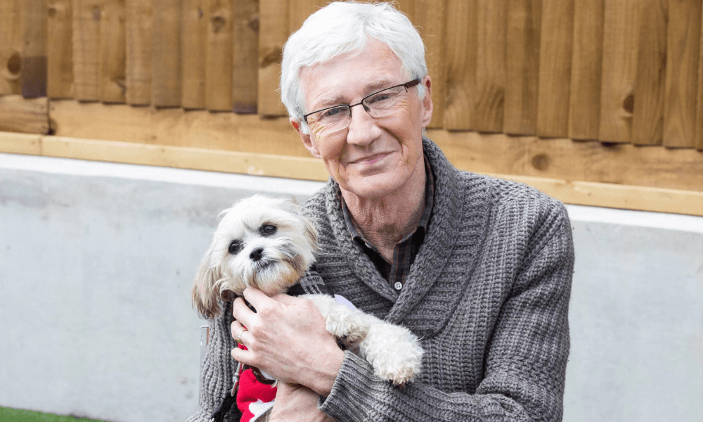 A senior man holding a small dog in a backyard setting.