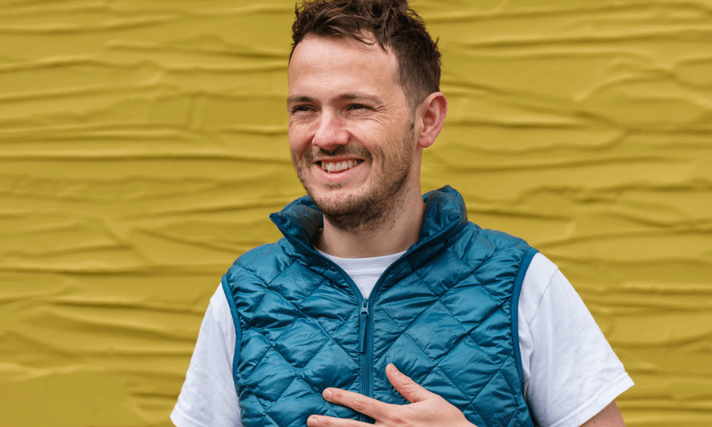Man smiling in a blue vest against a yellow background.