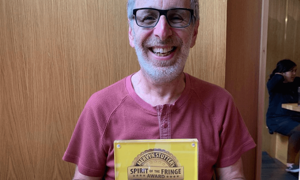 Smiling man sitting at a table holding a 'spirit of the fringe' award plaque.