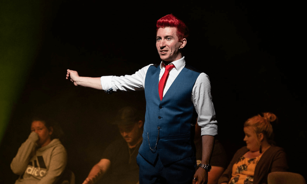 A person with red hair wearing a vest and tie gestures on stage with an audience in the background.