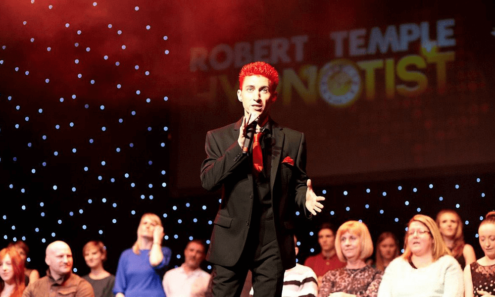 A performer with red hair on stage during a hypnosis show with an audience in the background.