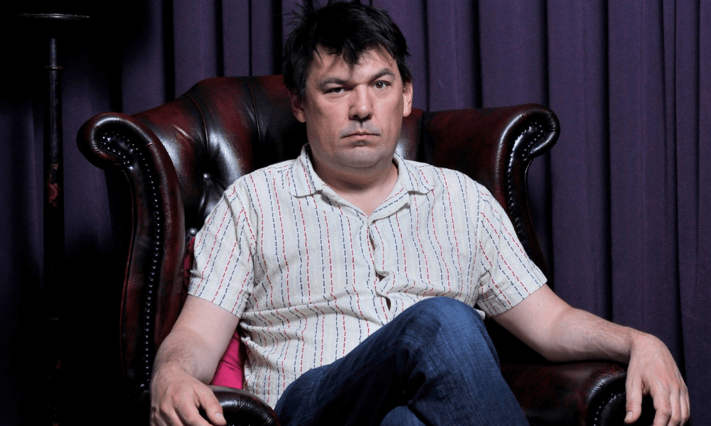 A man sitting in an armchair with a serious expression in front of purple curtains.