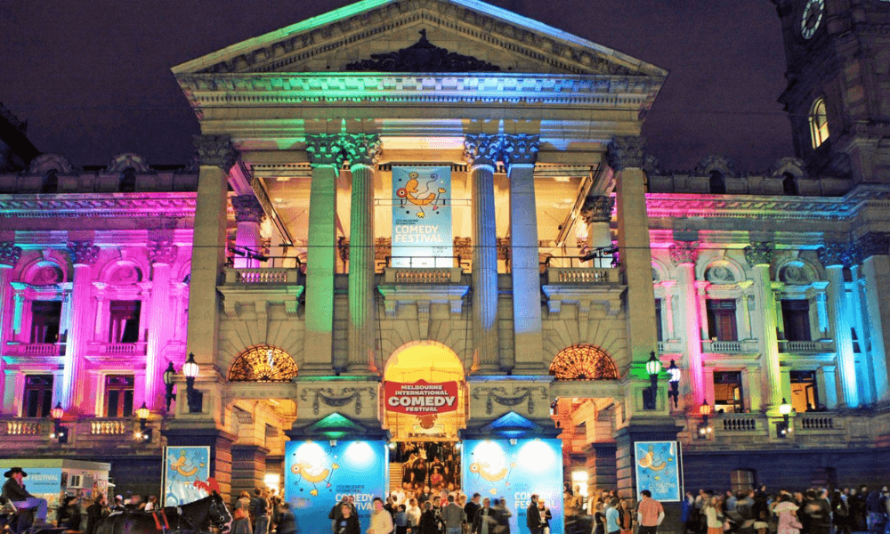 A vibrant evening scene at a comedy festival with colorful lights illuminating a historic building facade as attendees gather at the entrance.