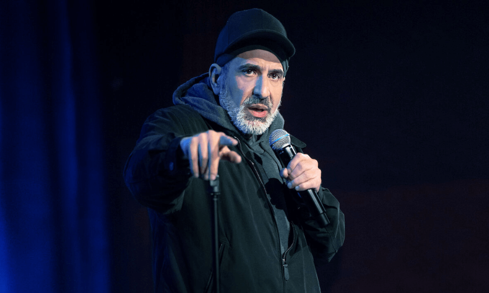 Comedian on stage performing with a microphone, pointing towards the audience.
