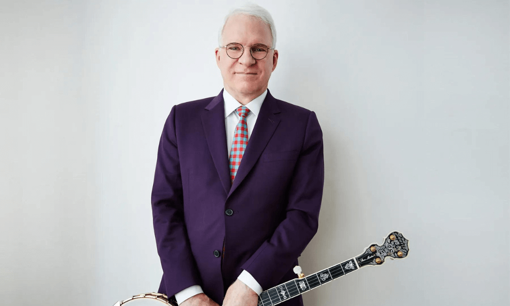 A man in a purple suit holding a banjo stands against a light background.