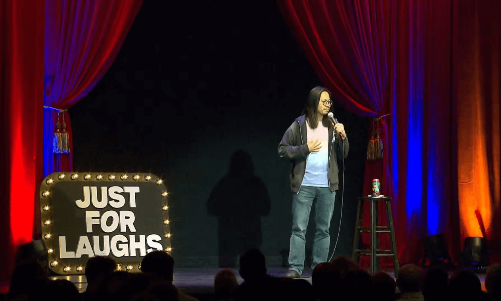 A man standing on stage with a sign that says just for laughs.