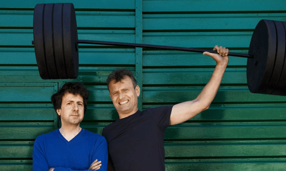 Two men posing with a barbell in front of a green wall.