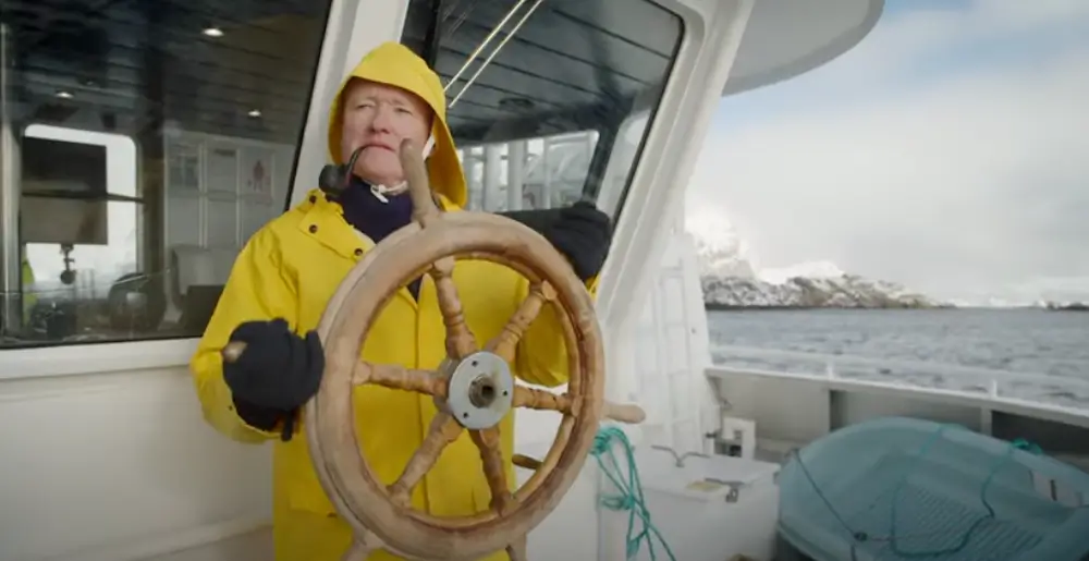 A person in a yellow raincoat steering a boat's wheel with a thoughtful expression.