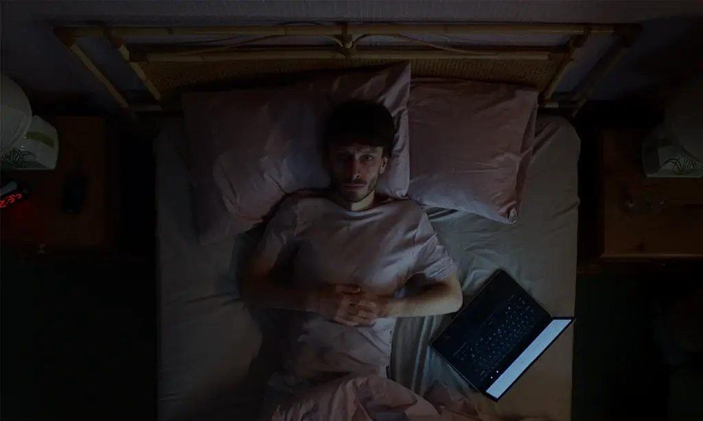 Man lying in bed and looking up at the camera, with a laptop open beside him, in a dimly lit room.