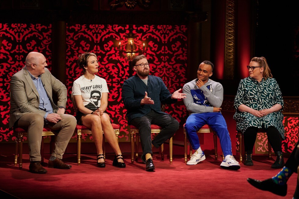 Five people sitting in a row on stage, engaged in a discussion, with an ornate red and gold background in a theater setting.