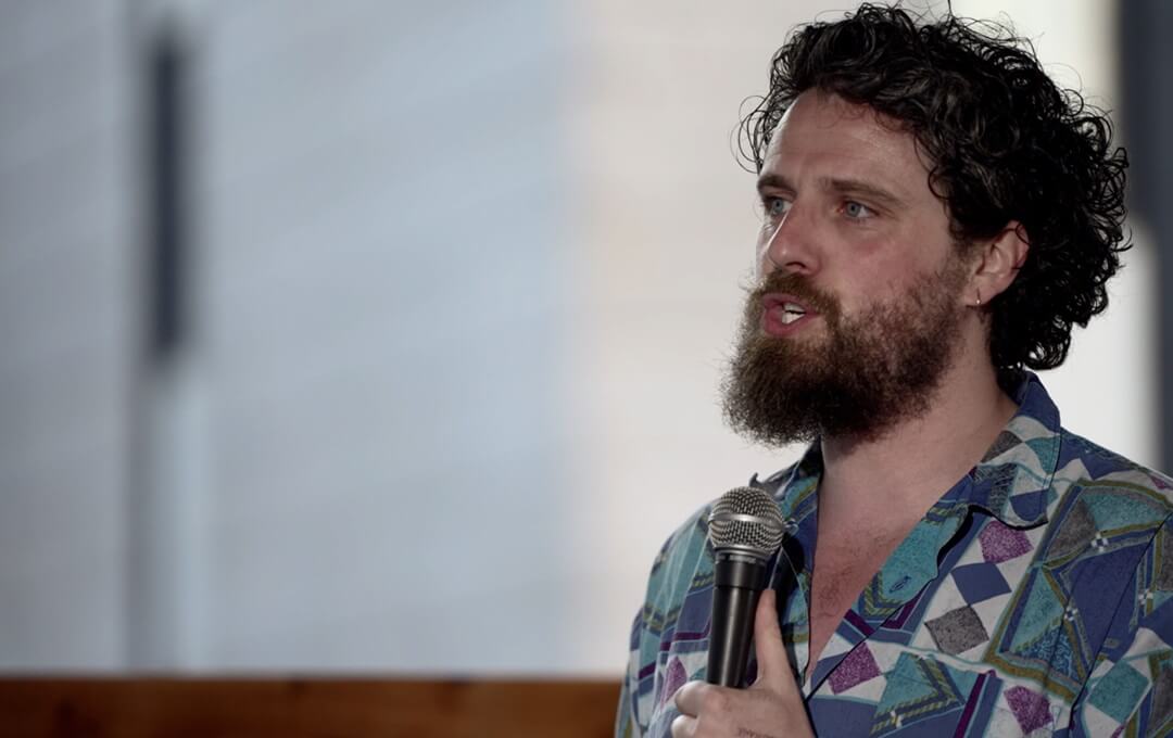 A man with curly hair and a beard speaking into a microphone, wearing a colorful patterned shirt.
