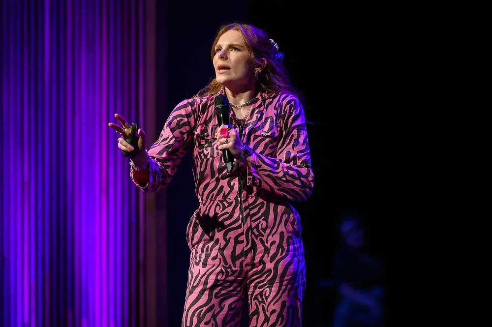 A person in a zebra-striped outfit speaks into a microphone on a stage with purple lighting.