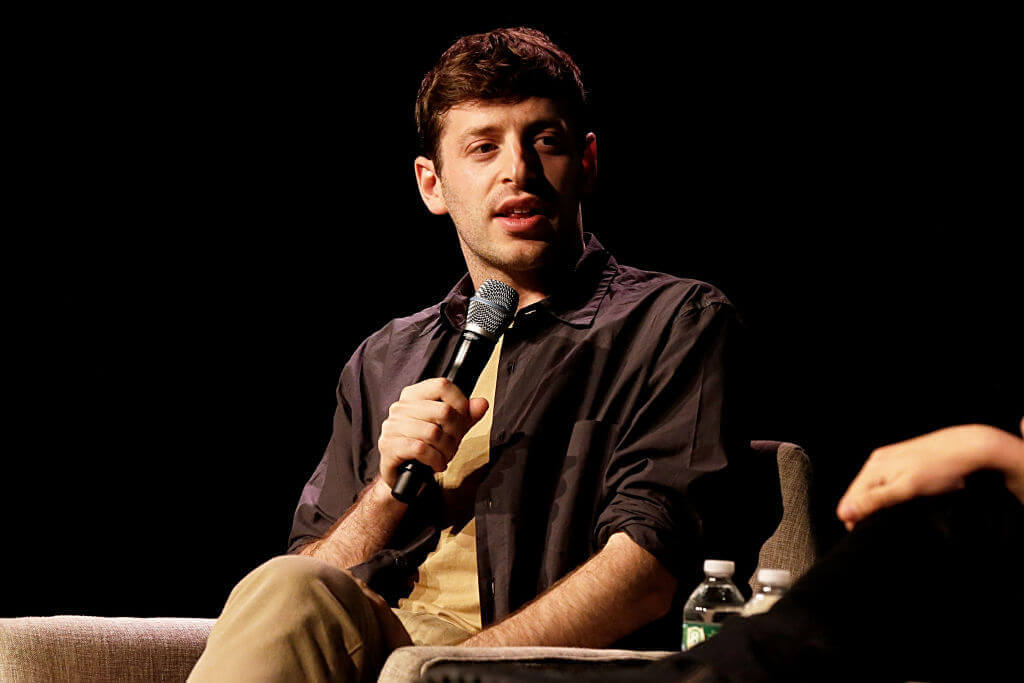 A young man speaks into a microphone during a discussion on stage, wearing a dark shirt and khaki pants.