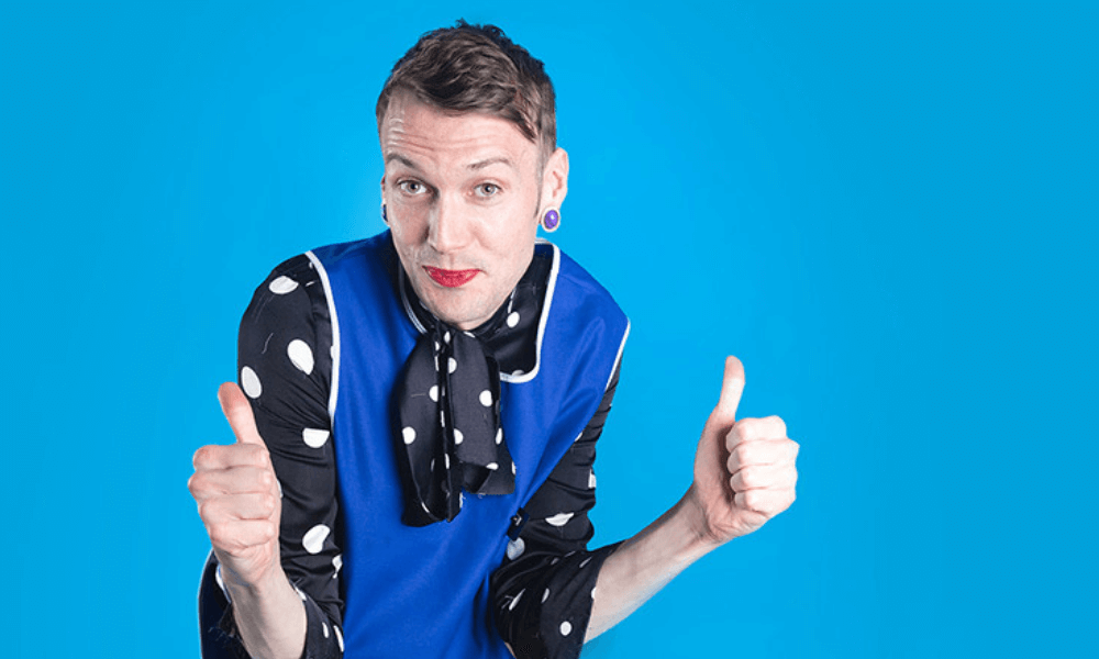 Man in polka dot shirt and scarf giving two thumbs up against a blue background.