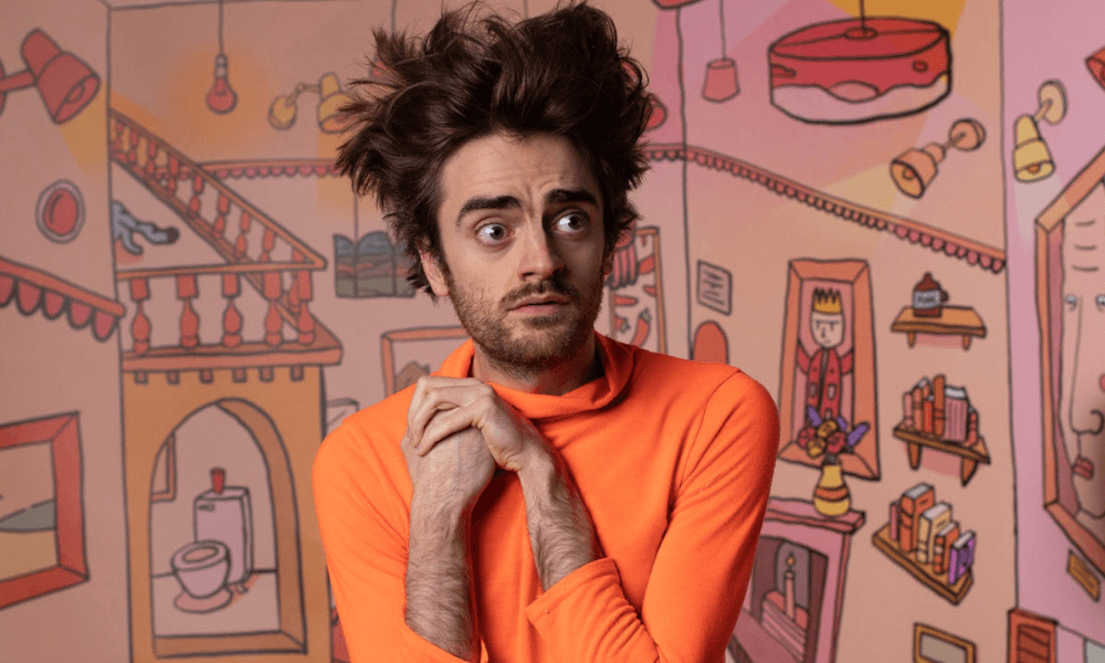 A man with messy hair and a surprised expression wears an orange turtleneck, posing in front of a whimsical cartoon-style wall mural.