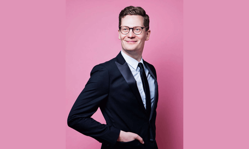 A man in a suit with glasses smiling confidently against a pink background.