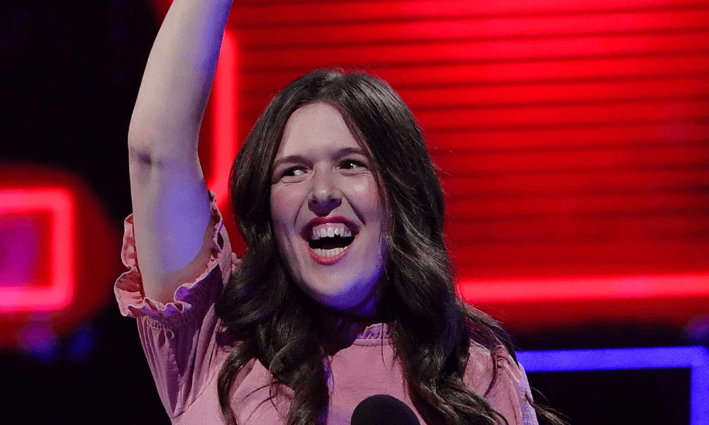 A woman with long brown hair, wearing a pink blouse, smiling broadly and raising her right arm in a victory gesture on a stage with a red background.