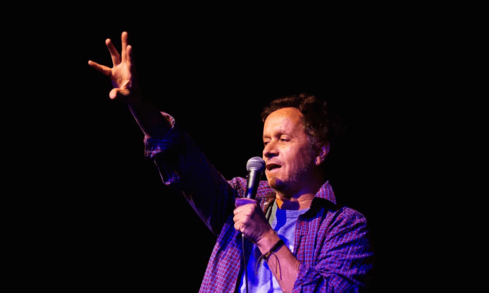 A person on stage speaking into a microphone with one hand raised expressively.