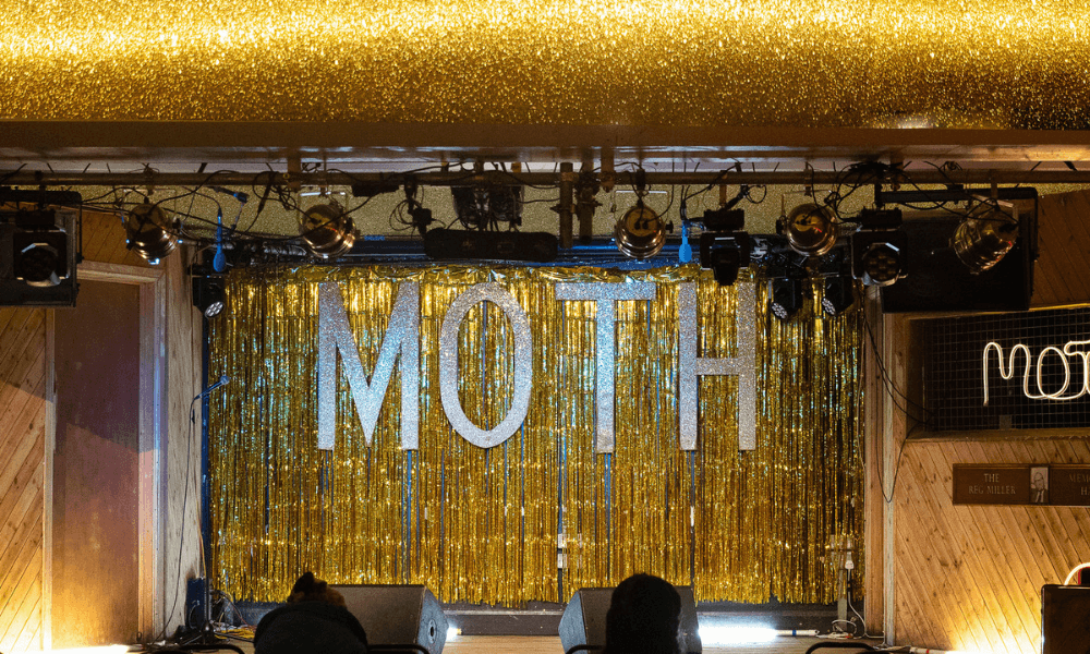 A stage with golden curtains and the word "moth" prominently displayed, with stage lights and speakers on either side.