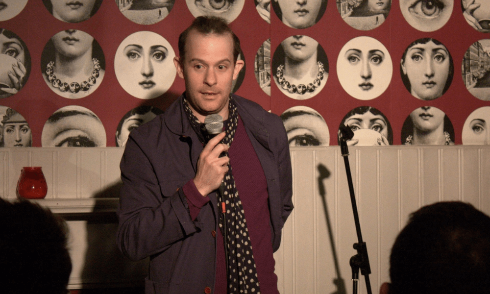 A man performing stand-up comedy in front of a wallpaper adorned with vintage faces.