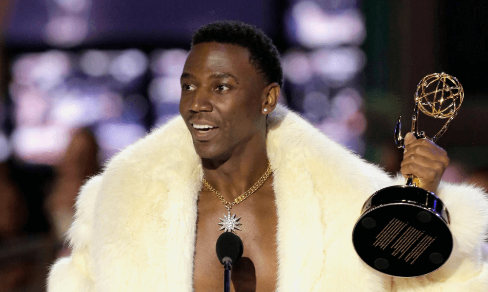 A person wearing a white fur coat and a gold chain, holding an emmy award on stage.
