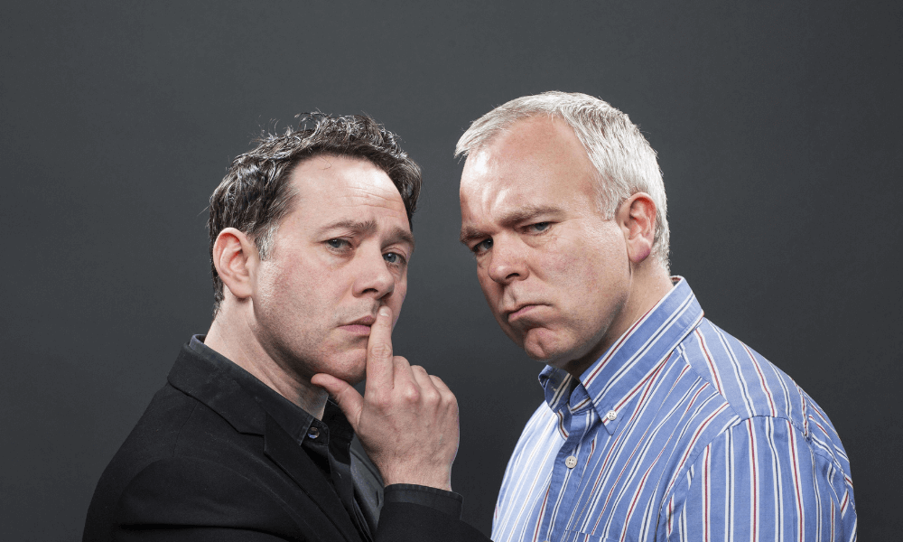 Two men facing the camera, one placing his finger to his lips in a silencing gesture, with serious expressions on a gray background.
