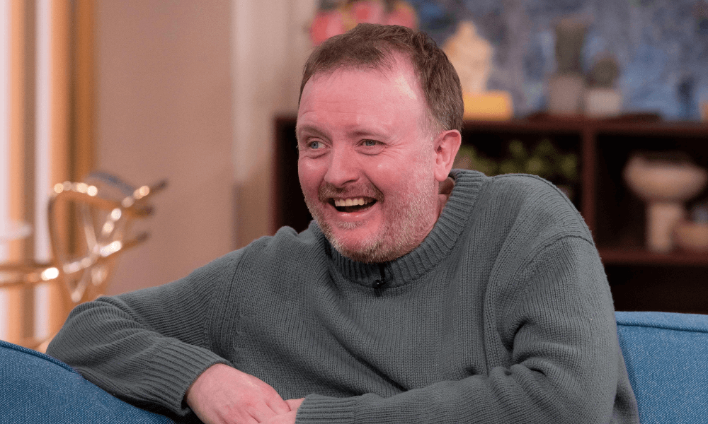 Man in green sweater smiling while seated on a blue couch.