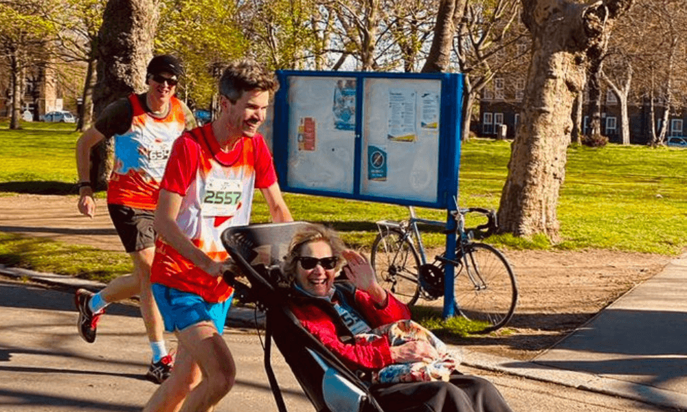 Two runners, one pushing a companion in a wheelchair, participating in a race on a sunny day in a park.