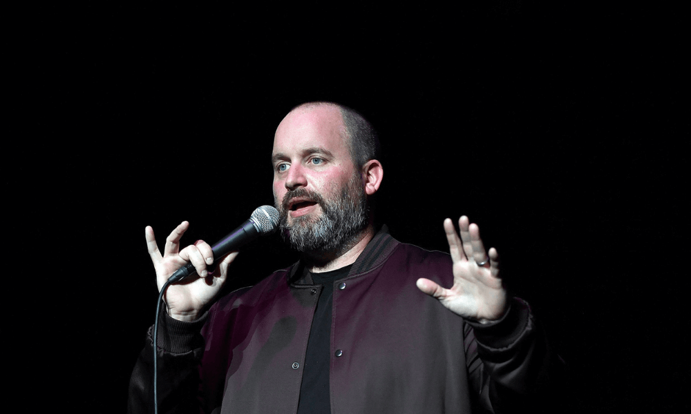 A man with a beard speaking into a microphone on a dark stage, gesturing with his hands.