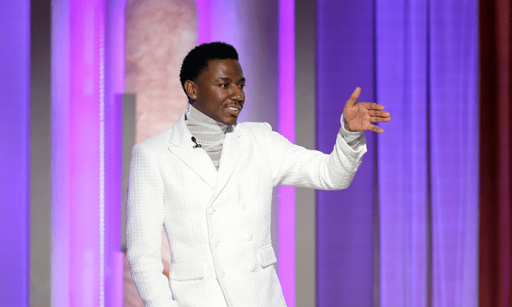 A man in a white suit and turtleneck smiling and gesturing on a stage with colorful vertical lights in the background.