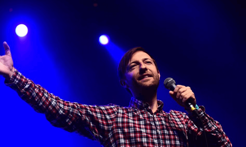 A man in a plaid shirt smiling and gesturing with one hand while holding a microphone on stage under blue lights.