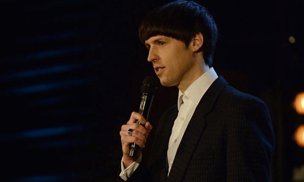 A man with a microphone stands on stage giving a speech, dressed in a dark suit and sporting a bowl haircut.