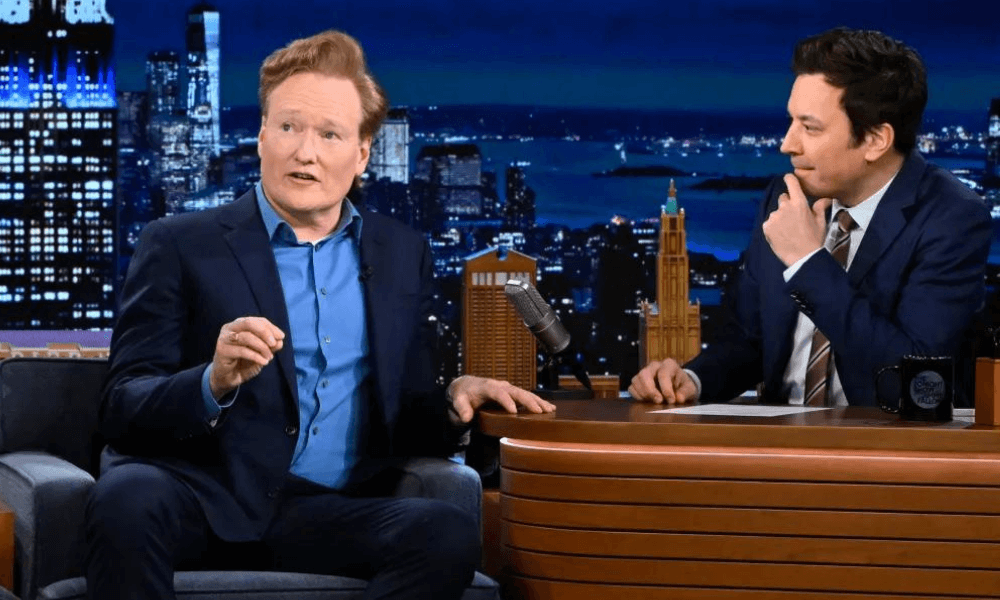 Two men conversing on a late-night talk show set; one gestures while speaking and the other listens attentively, with a cityscape background.