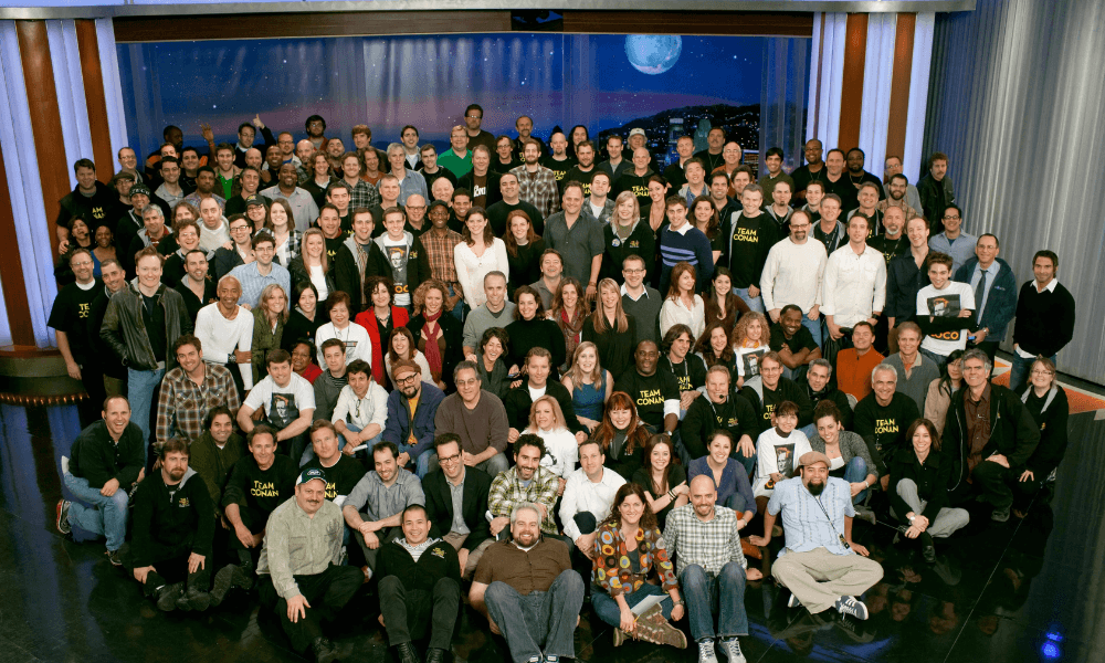 Large group of people posing for a photo in a room with a night sky background. some are standing, others seated, diverse ages and attire, smiling at the camera.